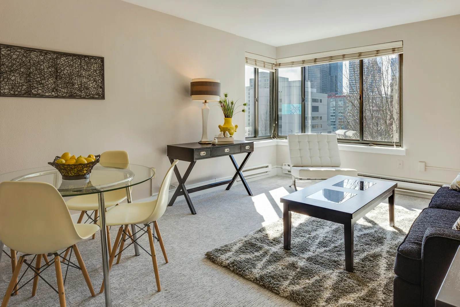 Reference photograph: a compact living room with a glass coffee table, pale chairs and large windows.